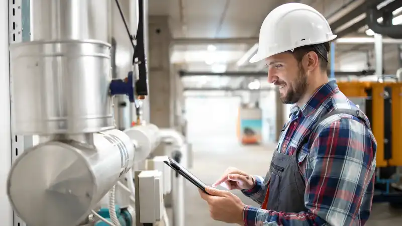 Engineer in a hard hat inspecting industrial pipes with a tablet.